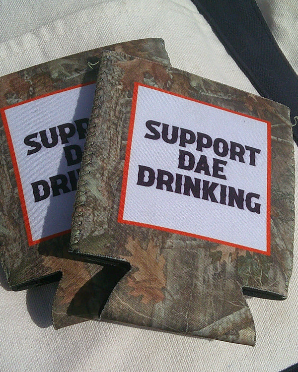 Camouflage can coolers with 'Support Dae Drinking' text on a light fabric background