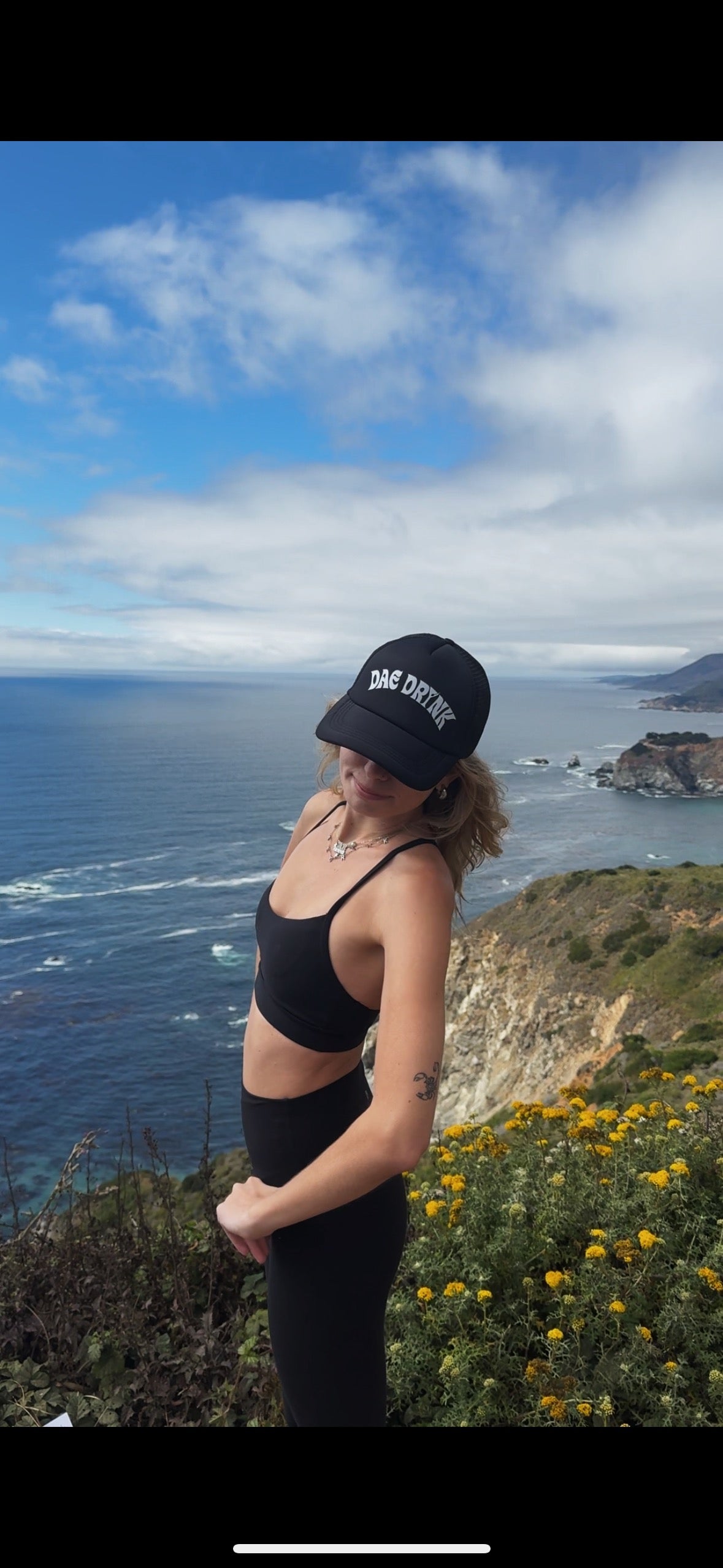 Person wearing a black cap and outfit standing on a cliff overlooking the ocean.